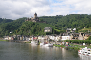 Ein malerischer Blick auf den Rhein in Deutschland mit einer Burg auf einem Hügel im Hintergrund, Booten auf dem Fluss, Fahrzeugen auf einer näheren Straße und einem bewölkten Himmel.