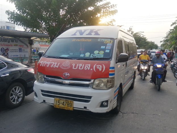 Ein weißer Lieferwagen fährt eine Straße mit parkenden Autos und Motorrädern entlang, mit Menschen in Helmen auf den Fahrzeugen, Bäumen, Gebäuden, Texttafeln, Polen, Drähten und einem klaren blauen Himmel im Hintergrund.
