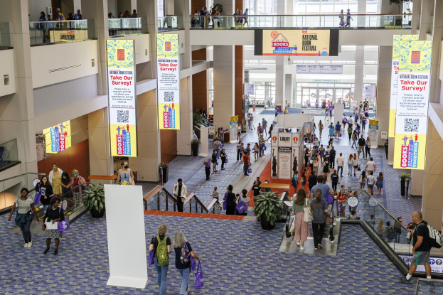 Eine große Gruppe von Menschen geht durch ein großes Gebäude mit Bannern, Topfpflanzen, Rolltreppen, Geländern, Säulen, Texttafeln, Glastüren und anderen Gegenständen, einige tragen Taschen, wahrscheinlich nehmen sie am Nationalen Buchfestival teil.