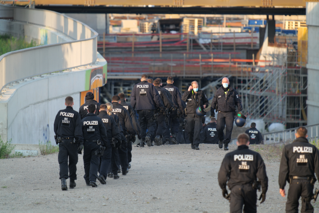 Eine Gruppe von Polizeibeamten geht auf einem Schotterweg, mit einer Wand auf der linken Seite und Pflanzen auf der rechten Seite, eine Brücke und andere Gegenstände im Hintergrund.