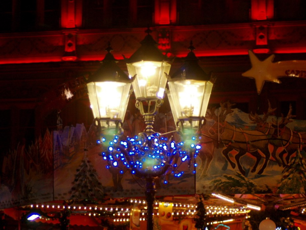 Eine festlich geschmückte Straßenlaterne mit Lichtern und Dekorationen steht nachts in einer Stadt, mit einem Gebäude im Hintergrund.