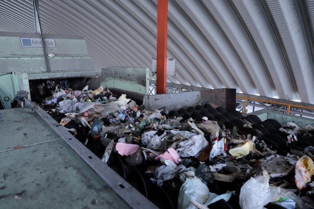 Großer Haufen von Mischabfällen auf einem Förderband in einer Recyclinganlage mit Maschinen und einem Metalldach darüber.