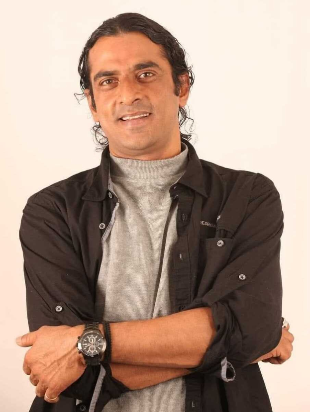 Ein Mann in einem schwarzen und aschfarbenen Anzug mit verschränkten Armen, der eine Uhr trägt, steht vor einem weißen Hintergrund und sieht nachdenklich aus.