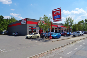 Ein Autohaus mit mehreren geparkten Autos davor, umgeben von Gebäuden, Bäumen, Strommasten, Schildern und einer Straße unter einem bewölkten Himmel.