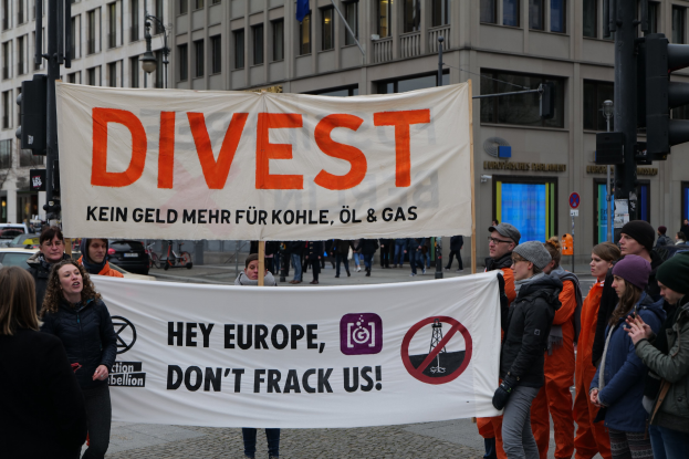 Gruppe von Menschen mit einem Banner, auf dem "Divest Hey Europe, Don't Frack Us" steht, auf einer Straße mit Gebäuden, Straßeninfrastruktur, Fahrzeugen, Bäumen und einem klaren Himmel im Hintergrund.