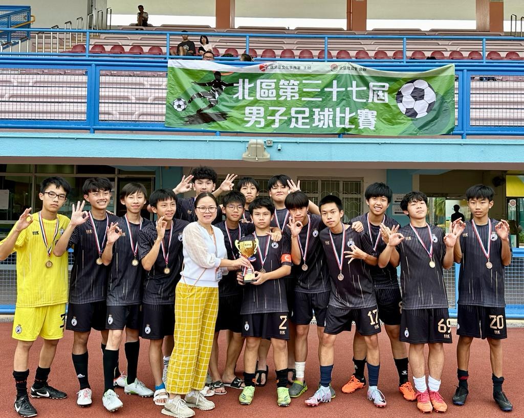 Gruppe junger Männer in Fußballtrikots auf einem Feld, die Medaillen tragen und einen Pokal halten, mit einem "Yokohama U-16 Boys Soccer Team"-Schild im Hintergrund und Zuschauern in der Nähe.