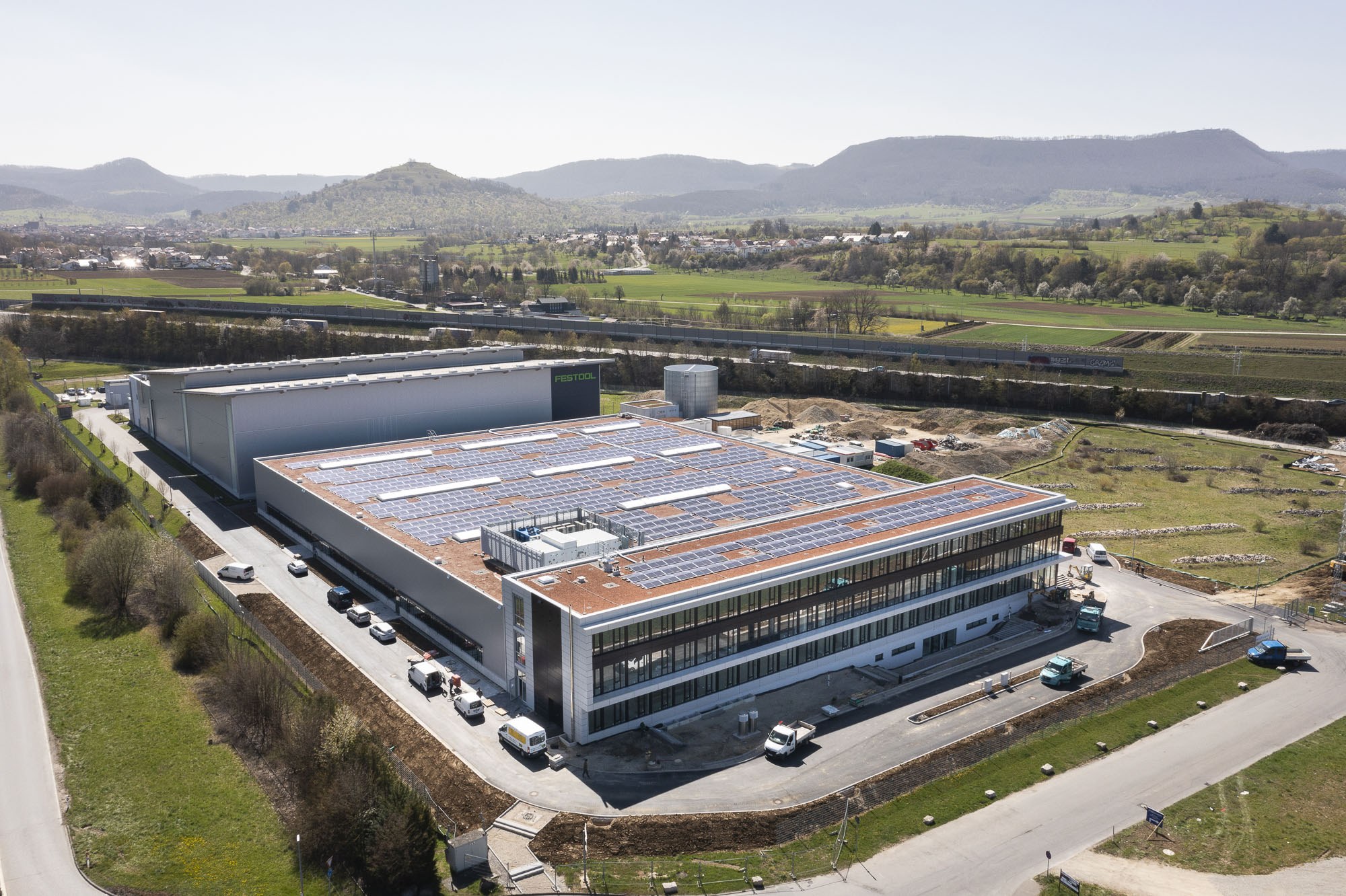 Luftaufnahme eines großen Gebäudes mit Solarmodulen auf dem Dach, umgeben von Fahrzeugen, Bäumen, Gras und Masten, mit Hügeln und einem klaren blauen Himmel im Hintergrund; das Gebäude ist Deutschlands erste solarbetriebene Fabrik.