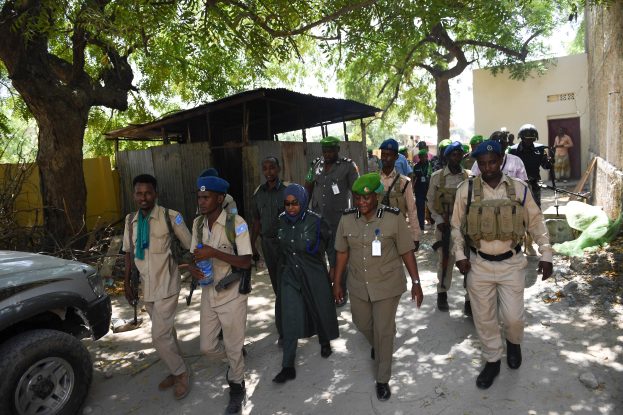 Eine Gruppe von Polizisten in Uniform und Mützen, einige mit Waffen, die eine Straße entlanggehen neben einem Auto mit Bäumen, Schuppen, Wänden und Gebäuden im Hintergrund unter einem sichtbaren Himmel.