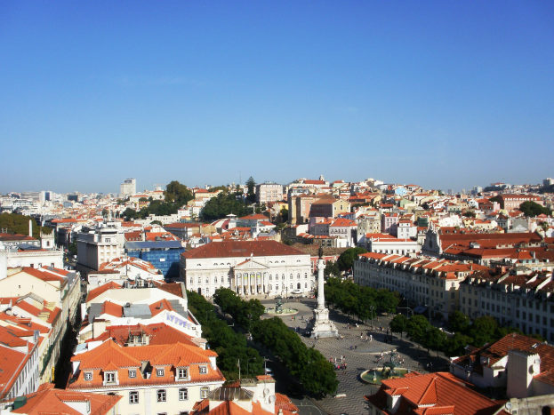Ausblick auf Lissabon von einem Hügel mit Gebäuden, Bäumen, einer Statue auf einem Sockel, Menschen auf einer Straße und dem Himmel im Hintergrund.