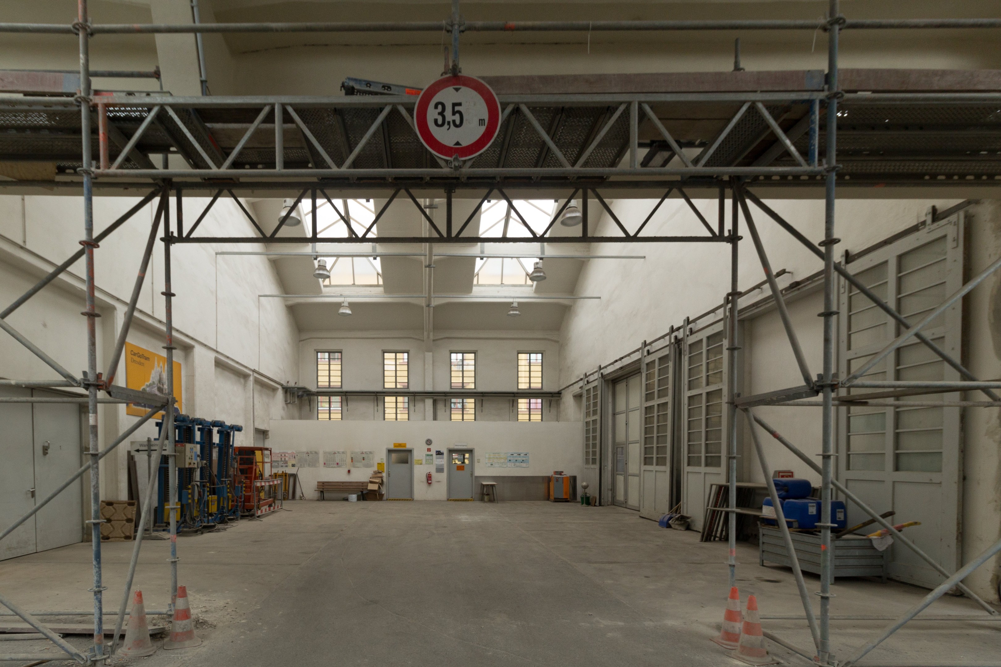 Innenansicht eines großen Lagerhauses mit ausgedehnten Gerüsten, Verkehrskegeln, Metallstangen, einem Schild, Maschinen, einer Tür, Fenstern, Wänden, Deckenlampen und einem Dach.