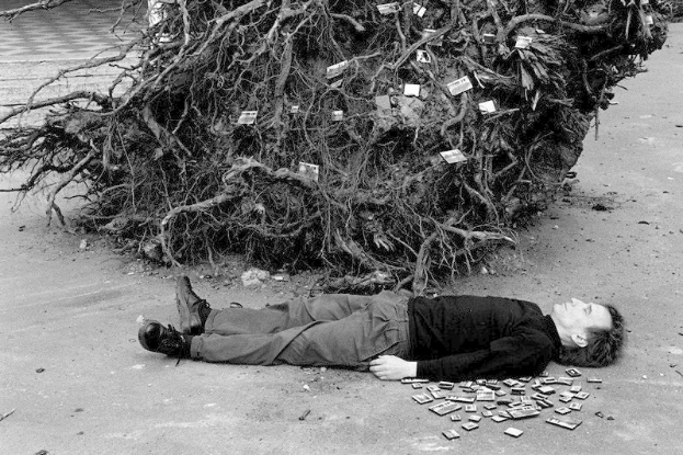 Mann in Wanderschuhen liegt neben Geldhaufen auf dem Boden, mit Baum im Hintergrund, auf Schwarz-Weiß-Foto.