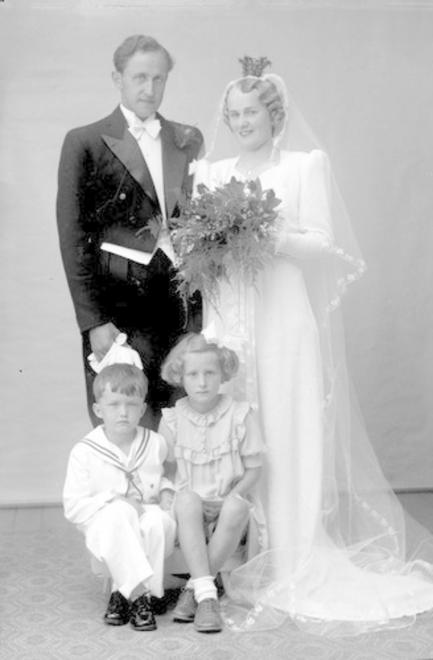 Schwarz-weiß-Fotografie einer Braut in einem weißen Kleid, die einen Strauß hält, ein strahlender Bräutigam neben ihr und zwei Kinder, die vor ihnen auf dem Boden sitzen.