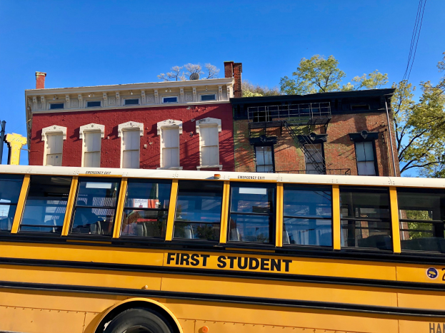 Gelber Schulbus mit der Aufschrift "First Student" vor einem roten Backsteingebäude geparkt, mit Menschen im Inneren, Bäumen und einem klaren blauen Himmel im Hintergrund.