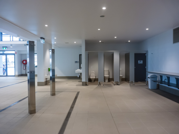 Große Öffentliche Toilette mit mehreren Toiletten, tragenden Säulen, einem Mülleimer, einer Eingangstür, einem Schild, einem Feuerlöscher und Deckenleuchten.