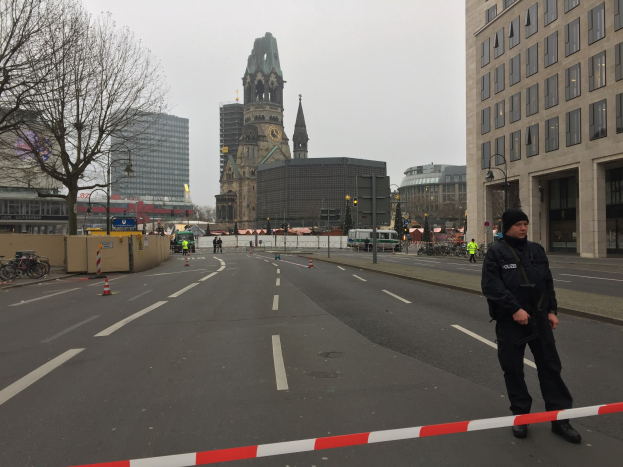 Polizeibeamter in schwarzer Uniform und Mütze auf einer Berliner Straße umgeben von Absperrbaken, Fahrrädern, Laternenmasten, Bäumen und Gebäuden mit einem Kirchturm und einem klaren blauen Himmel im Hintergrund.