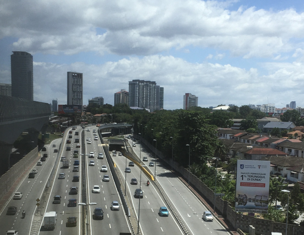 Eine vielbefahrene Autobahn mit hohen Gebäuden, eine Brücke auf der linken Seite, Laternenpfählen, Bäumen und einem bewölkten Himmel im Hintergrund.