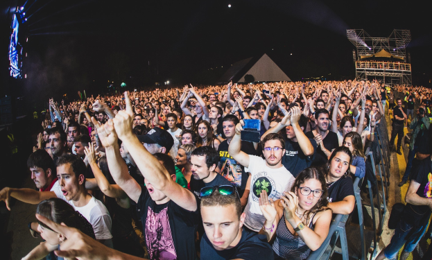 Menschenmenge auf einem Musikfestival mit erhobenen Händen, einige sitzen und andere stehen, begrenzt von einem Metallzaun, mit Gebäuden und Lichtern im Hintergrund unter einem dunklen Himmel.