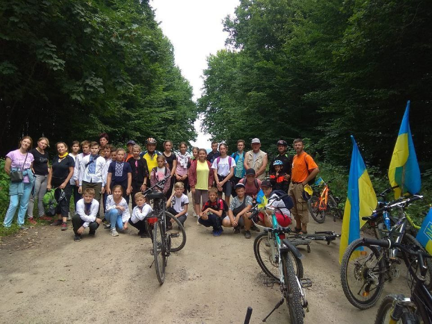 Eine Gruppe von Menschen steht neben Fahrrädern auf einem Schotterweg mit Bäumen und einem klaren blauen Himmel, einige tragen Helme und Taschen.