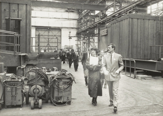 Eine Gruppe von Menschen in Anzügen geht durch eine Fabrik mit Maschinen auf der linken Seite und einer Wand im Hintergrund.