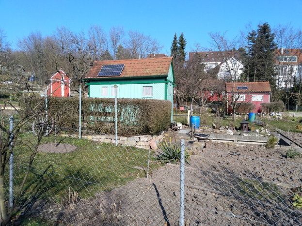 Kleines grünes Haus mit Solarpanelen auf dem Dach, umgeben von Maschendrahtzaun, vor einem klaren blauen Himmel mit Bäumen, Pflanzen und Gras im Vordergrund.