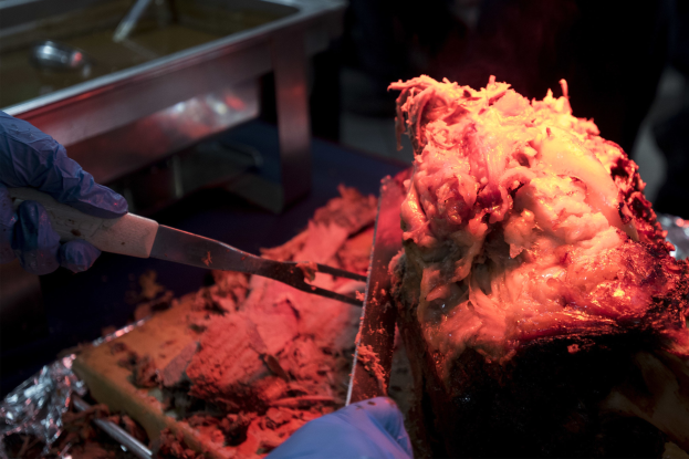 Eine Person mit Handschuhen hält ein Messer über ein Lebensmittel auf einem Tisch, mit Gegenständen im Hintergrund.