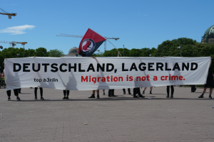 Eine Gruppe von Menschen mit einem Transparent, auf dem "Deutschland, Lagerland Migration ist kein Verbrechen" steht, und einer Fahne, die auf dem Boden steht, mit Straßenlaternen, Bäumen, Kränen, einem Gebäude und einem bewölkten Himmel im Hintergrund.