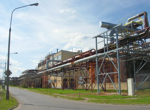 Großes Werk mit zahlreichen Rohren an seiner Außenseite, umgeben von Gebäuden, Straßeninfrastruktur, Grünflächen und einem bewölkten Himmel.