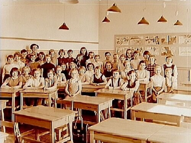 Ein Schwarz-Weiß-Foto eines Klassenzimmers mit Kindern auf Bänken und Stehplätzen, umgeben von Tischen und Stühlen, mit einer Tafel an der Wand und Deckenleuchten.