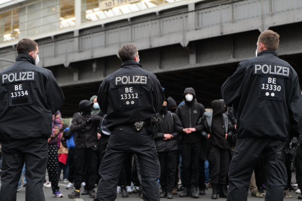 Polizeibeamte in schwarzen Uniformen und Masken stehen vor einer Menge bei einer Demonstration, mit einer Brücke und einem Gebäude im Hintergrund.