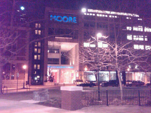 Ein Krankenhausgebäude mit parkenden Fahrzeugen davor, umgeben von Bäumen und einem Zaun.