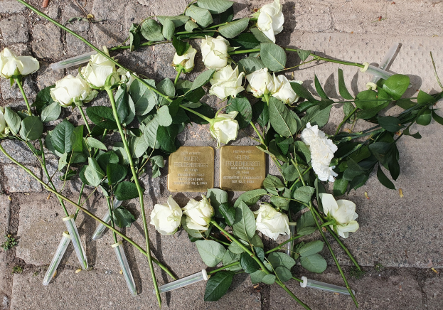 Ein Kränzchen aus weißen Rosen und grünen Blättern mit zwei goldfarbenen, beschrifteten Gegenständen, das auf dem Boden vor einem Berliner Denkmal liegt.
