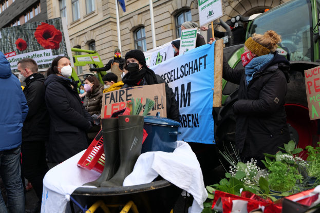 Eine Gruppe maskierter Demonstranten mit Schildern vor einem Lastwagen, mit einem Tisch, Pflanzen, Gebäuden und einer Fahne im Hintergrund, versammelte sich, um gegen ein Pestizidverbot zu protestieren.