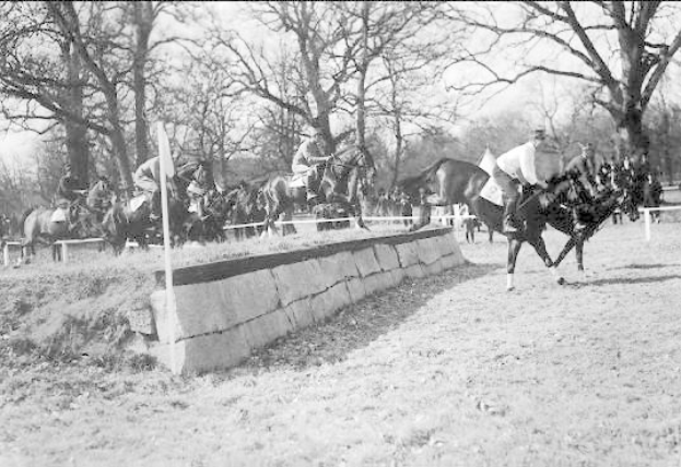 Schwarzes Foto von Menschen, die auf Pferden über eine grüne Wiese reiten, mit einer Mauer und Stangen im Vordergrund und Bäumen und Himmel im Hintergrund.