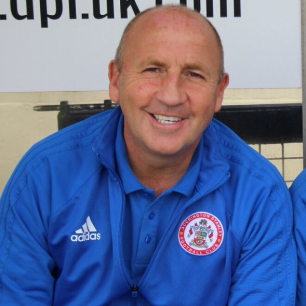 Zwei Männer in einem Raum, einer in einer blauen Jacke, der neben einem anderen Mann sitzt und lächelt, mit einer Tafel an der Wand dahinter.