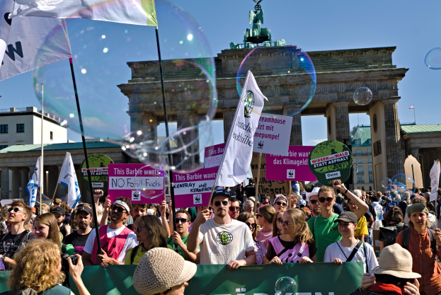 Eine Menschenmenge steht vor dem Brandenburger Tor in Berlin, viele tragen Mützen und Schutzbrillen, einige halten Fahnen und Transparente mit Text, mit Gebäuden, einem statuegekrönten Bogen und einem Himmel voller Seifenblasen im Hintergrund.