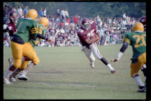 Fußballspieler in Bewegung mit dem Ball, umgeben von Gegenspielern und Mannschaftskameraden, mit Zuschauern und Bäumen im Hintergrund und Text unten.