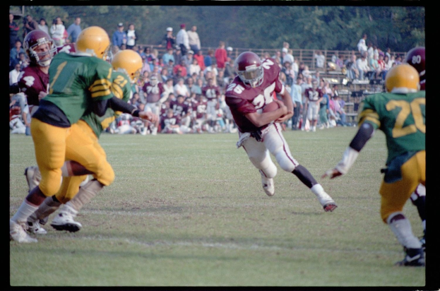 Fußballspieler in Bewegung mit dem Ball, umgeben von Gegenspielern und Mannschaftskameraden, mit Zuschauern und Bäumen im Hintergrund und Text unten.