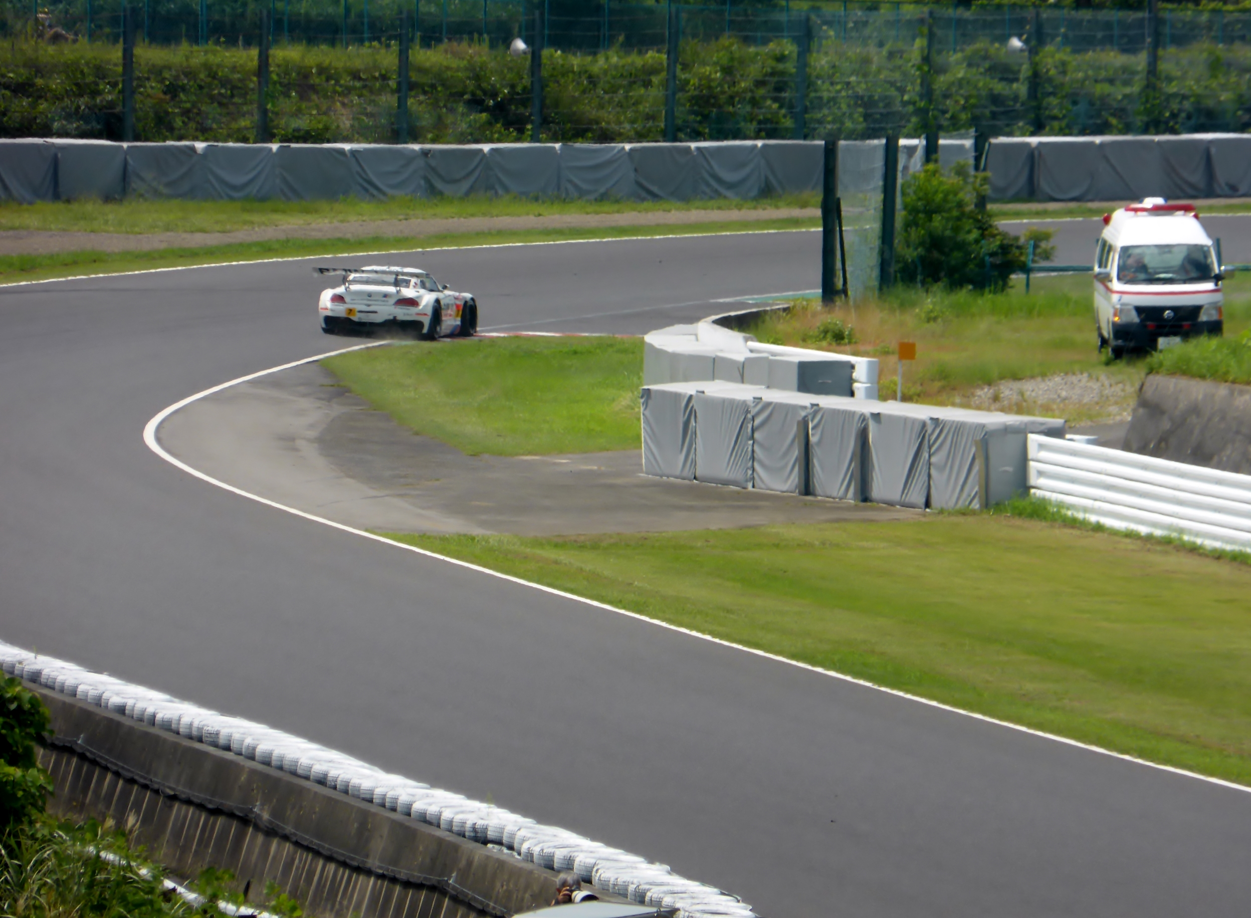 Ein Rennwagen fährt eine Rennstrecke entlang, im Hintergrund ist ein Polizeiauto zu sehen, umgeben von Gras, Pflanzen, Bäumen und Absperrungen, mit einem Fahrzeug auf der rechten Seite und Text unten.