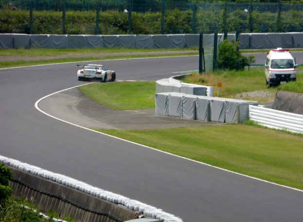 Ein Rennwagen fährt eine Rennstrecke entlang, im Hintergrund ist ein Polizeiauto zu sehen, umgeben von Gras, Pflanzen, Bäumen und Absperrungen, mit einem Fahrzeug auf der rechten Seite und Text unten.