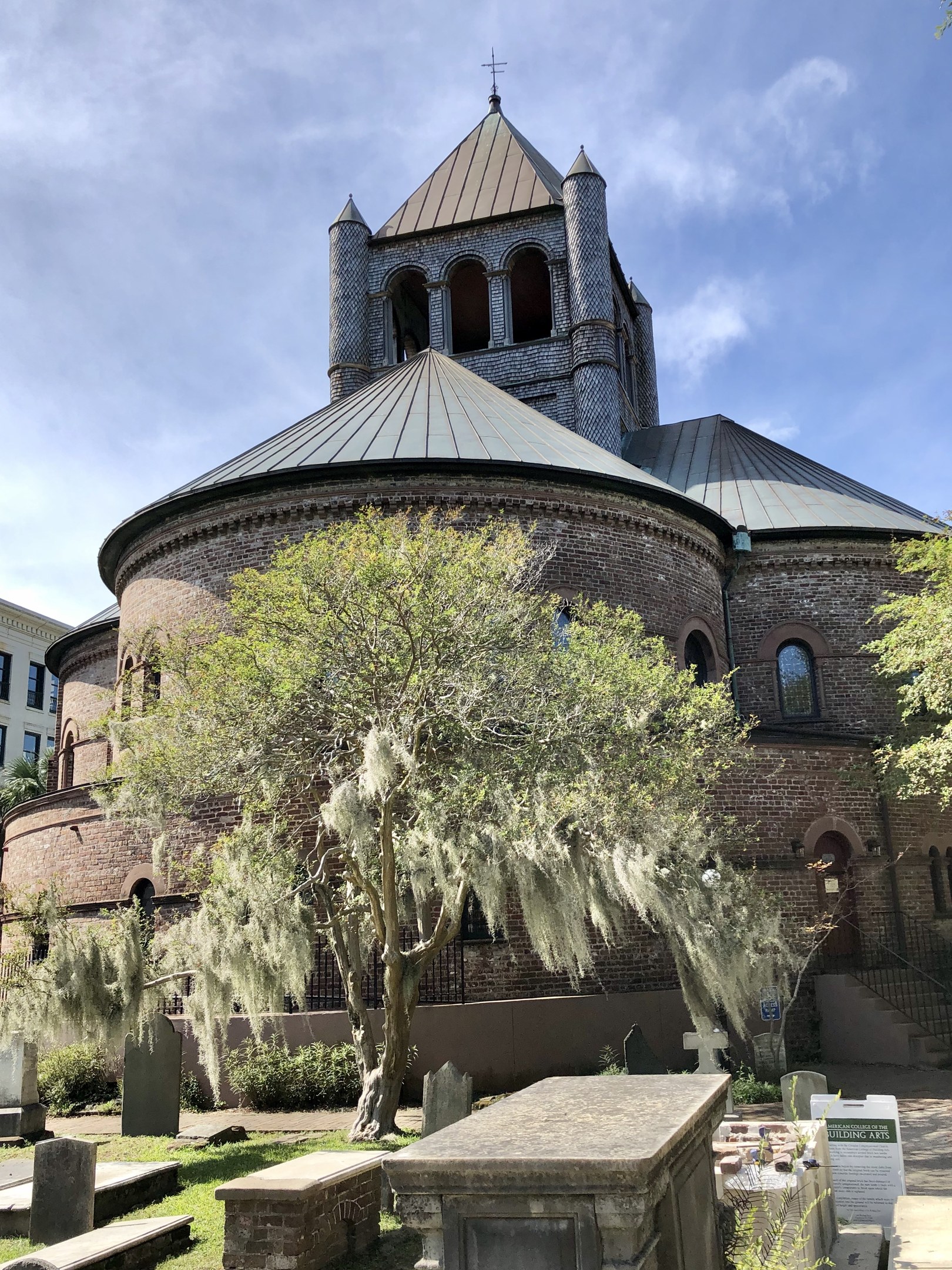 Außenansicht der Kirche des Heiligen Grabes in Charleston, South Carolina, mit einem Turm, einem Baum, umliegenden Gebäuden, einer Treppe und einem bewölkten Himmel, mit Gras und einer Texttafel unten.