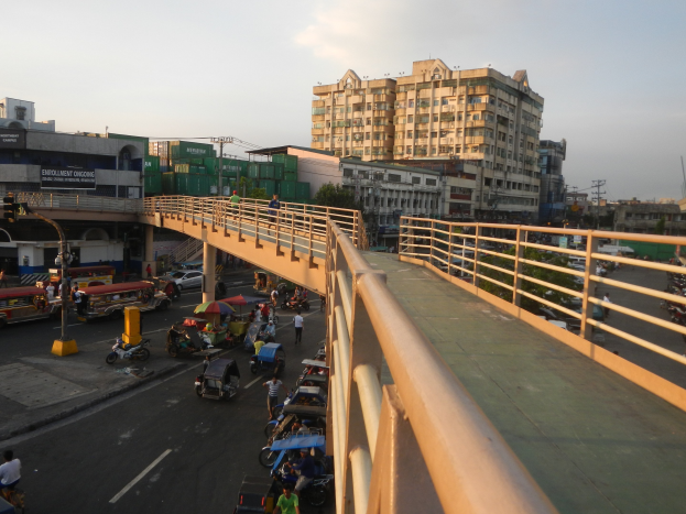 Eine Brücke spannt sich über eine vielbefahrene Straße mit Fahrzeugen, Fußgängern mit Regenschirmen und anderen Objekten darunter; Gebäude, Bäume, Pfosten und ein klarer blauer Himmel sind im Hintergrund zu sehen.