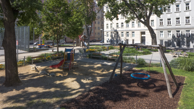 Ein Park mit einem Spielplatz in der Mitte, umgeben von Bäumen, Schaukeln, Bänken, Pflanzen, Gras, Steinen, einem Zaun, Pfosten, Fahrzeugen auf der Straße und Gebäuden mit Fenstern im Hintergrund.