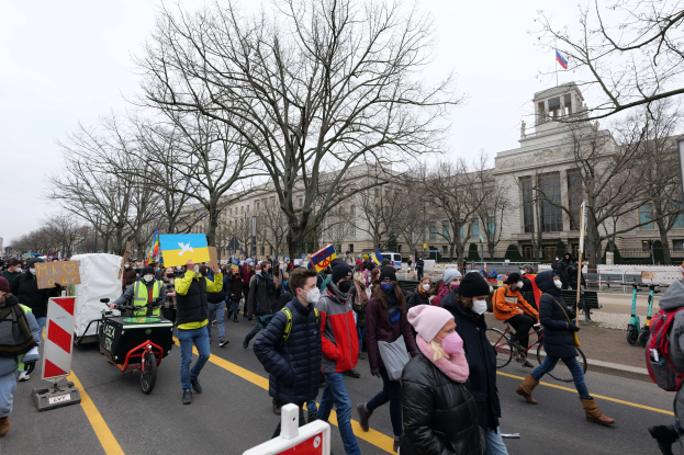 Eine große Gruppe von Menschen marschiert auf einer Straße in einer Demonstration, einige halten Schilder und andere fahren Fahrräder, mit Bäumen und einem Gebäude im Hintergrund unter einem klaren blauen Himmel.