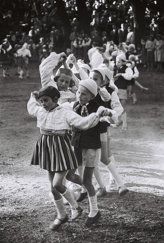 Schwarz-weißes Bild von Kindern, die in einem Park tanzen, mit einigen Zuschauern und Bäumen im Hintergrund.