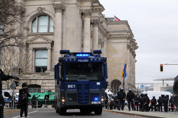 Gruppe von Polizeibeamten vor einem großen Gebäude mit architektonischen Details, Fahrzeuge auf der Straße, eine Person mit einer Kamera auf der linken Seite, Bäume, Verkehrsampeln, Fahnenmasten und einen klaren blauen Himmel.