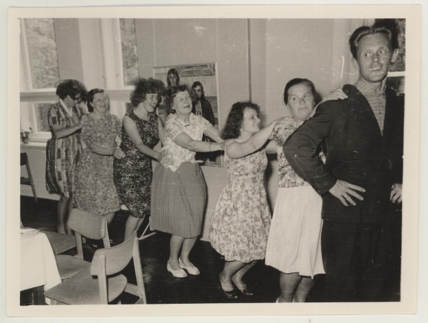 Gruppe von Menschen, die in einem Raum im Stil der 1950er Jahre tanzen, mit Stühlen, einem Tisch mit einer wei├čen Decke, einem Foto auf der Wand und Fenstern im Hintergrund.
