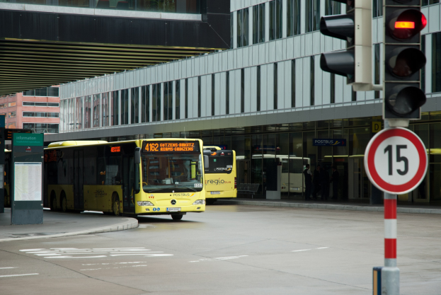 Zwei gelbe Busse fahren auf einer Straße mit hohen Gebäuden, ein Verkehrssignal mit einem Mast und einer daran befestigten Tafel auf der rechten Seite und einige Fußgänger im Hintergrund.