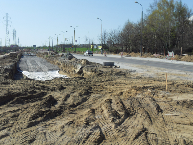 Großer Schutthaufen auf einer Baustelle neben einer Straße mit fahrenden Fahrzeugen, Laternenmasten, Strommasten mit Kabeln, Bäumen und einem klaren blauen Himmel im Hintergrund.