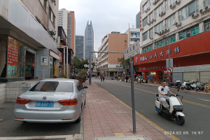 Ein Mann fährt auf einem Scooter eine Straße in einer Stadt mit hohen Gebäuden entlang, mit Menschen auf den Gehwegen und Fahrzeugen auf der Straße, unter einem klaren blauen Himmel mit Bäumen im Hintergrund.