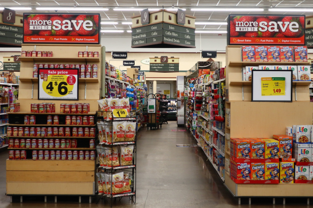 Ein Innenraum eines Lebensmittelladens mit verschiedenen Lebensmitteln auf Regalen, Ablagen und dem Boden, mit Deckenbeleuchtung und Informationsschildern, mit einer Person im Hintergrund.